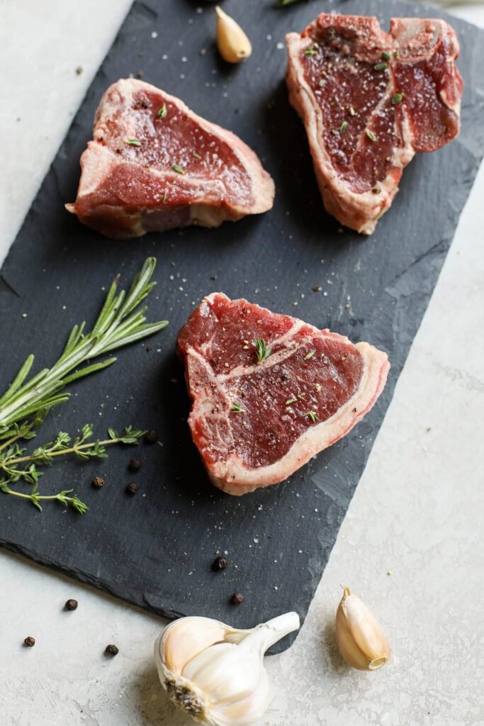 Raw lamb chops on black cutting board seasoned with salt and pepper and fresh herbs.