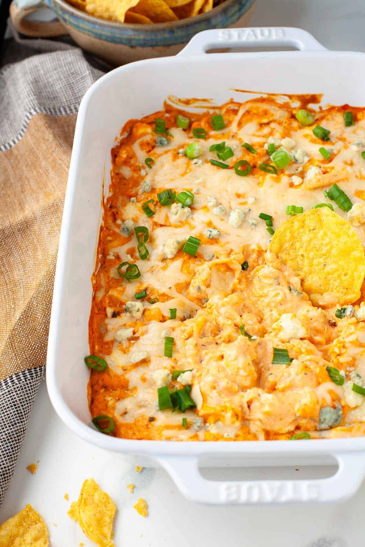 Buffalo Cauliflower Dip in baking dish garnished with sliced scallions and crumbled blue cheese with tortilla chip on top